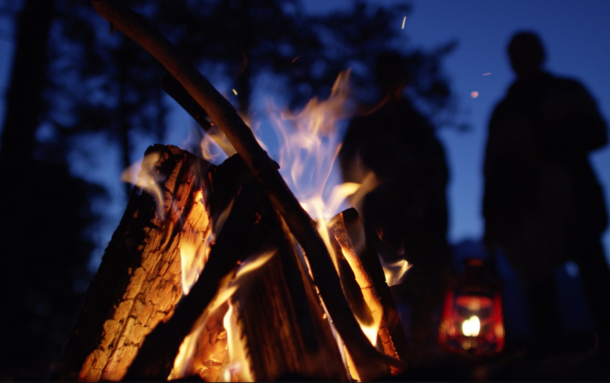 Campfire burning with two silhouetted figures at dusk