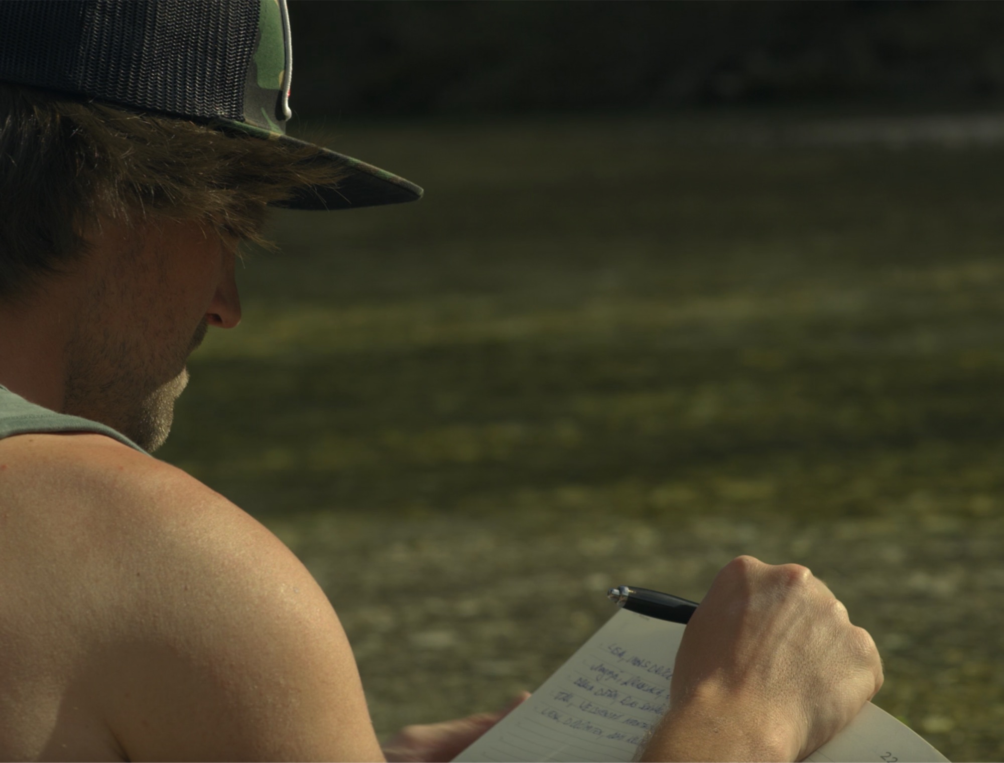 Man in cap journaling beside a sunlit river